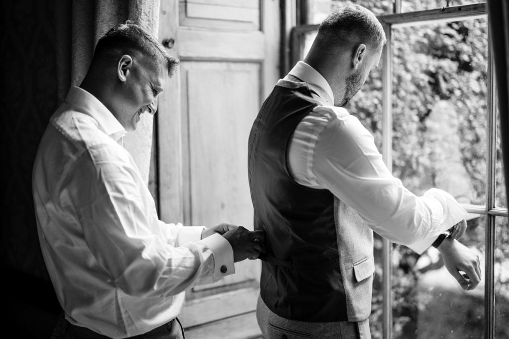 Man assisting groom with attire near window, monochrome photo.